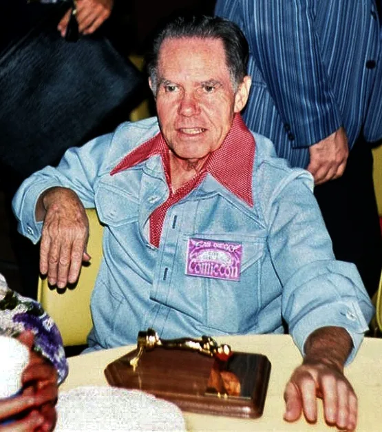 Daws Butler sits at a convention table wearing a blue shirt and a name badge.
