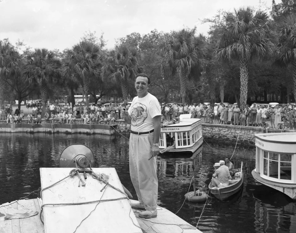 George Sidney stands on the bow of a boat with spectators and other boats behind him.