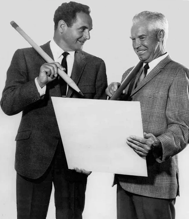 Black-and-white portrait of Joseph Barbera holding a large pencil beside William Hanna holding a board.