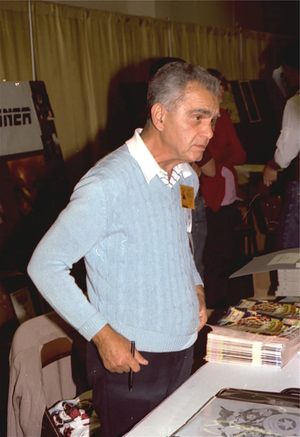 Jack Kirby stands at a convention table in a pale blue sweater.