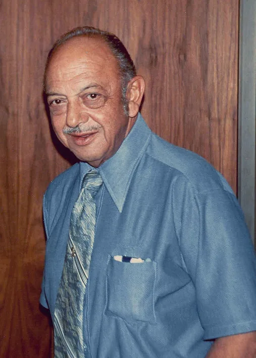 Mel Blanc stands smiling in a blue shirt and patterned tie in front of wood paneling.