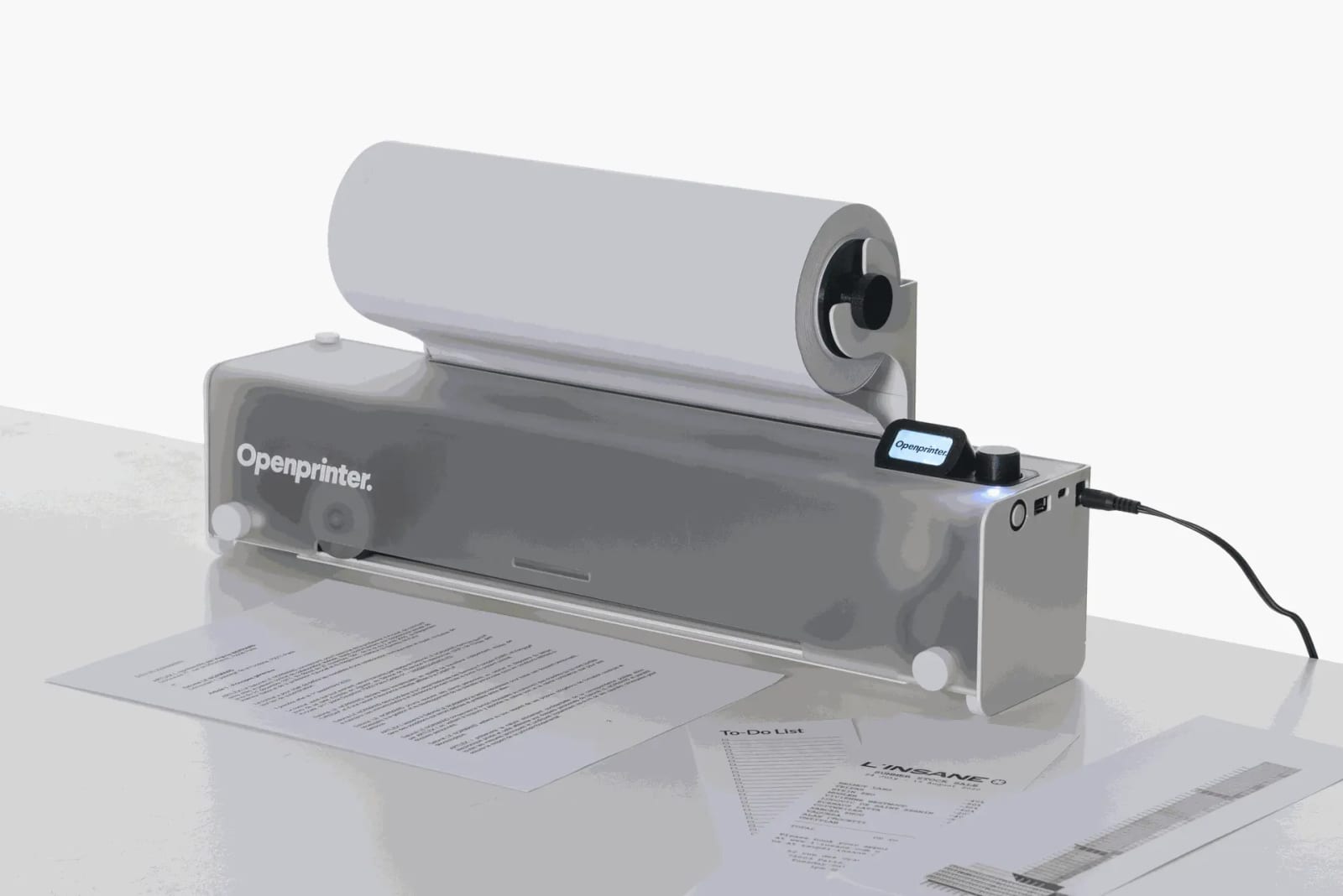 The grey Openprinter on a white desk with a paper roll on top, a printed contract page coming out of the front and a to-do list and shop flyer beside it