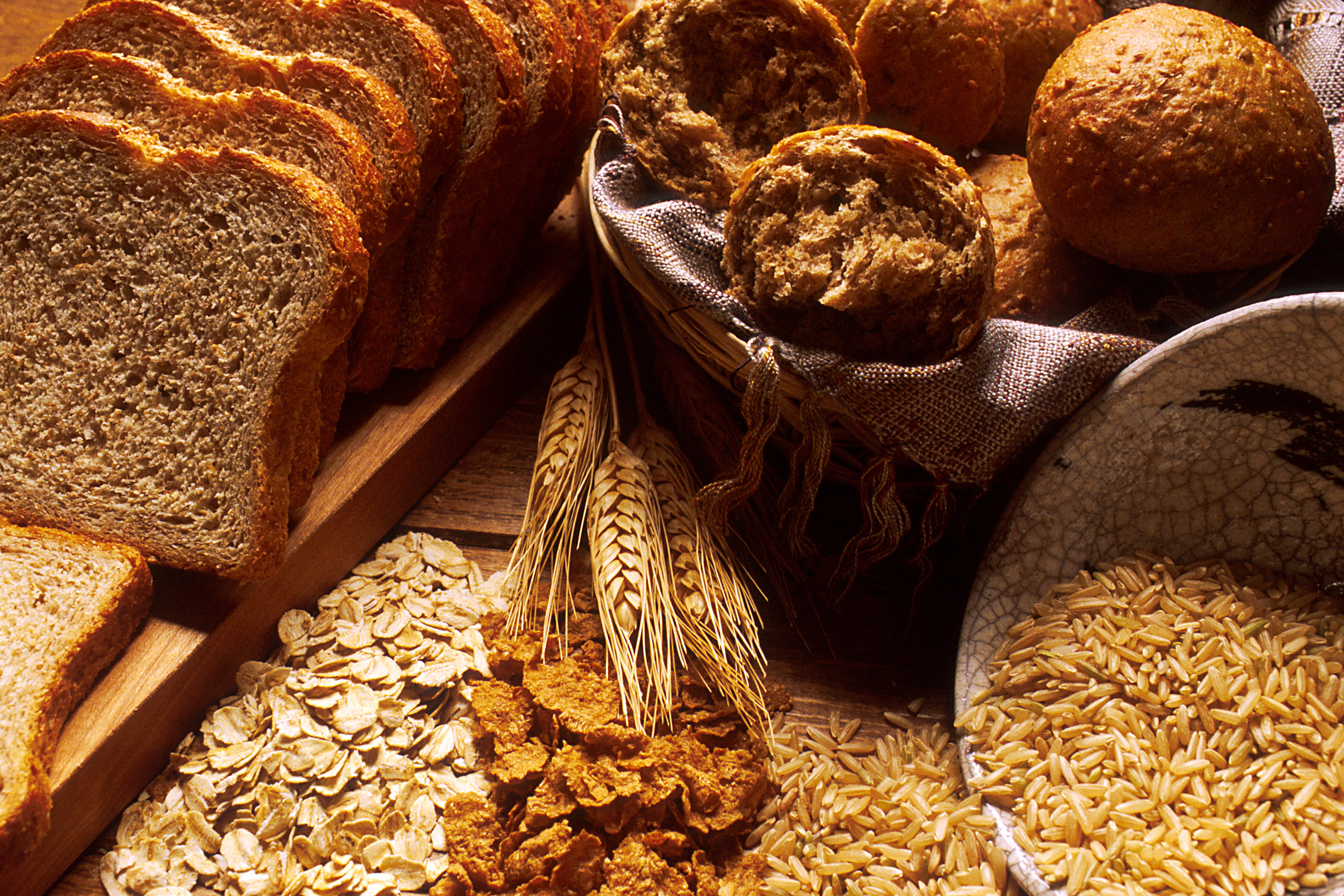 A sliced brown loaf, bread rolls and bowls of grains on a wooden table